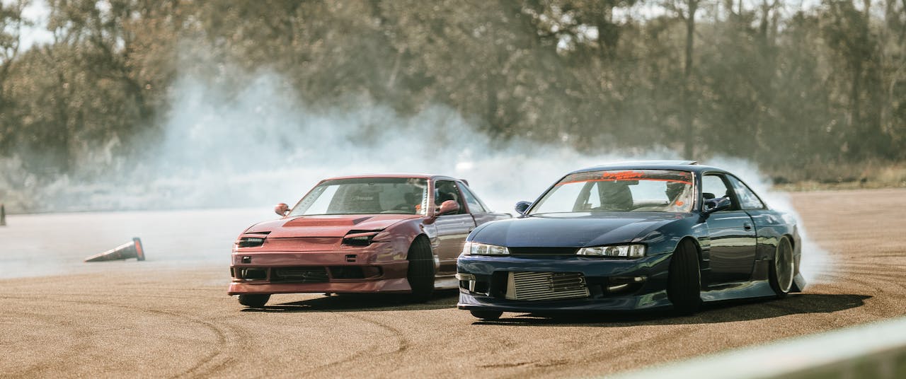 Two sports cars drift around a track emitting smoke, showcasing speed and precision.